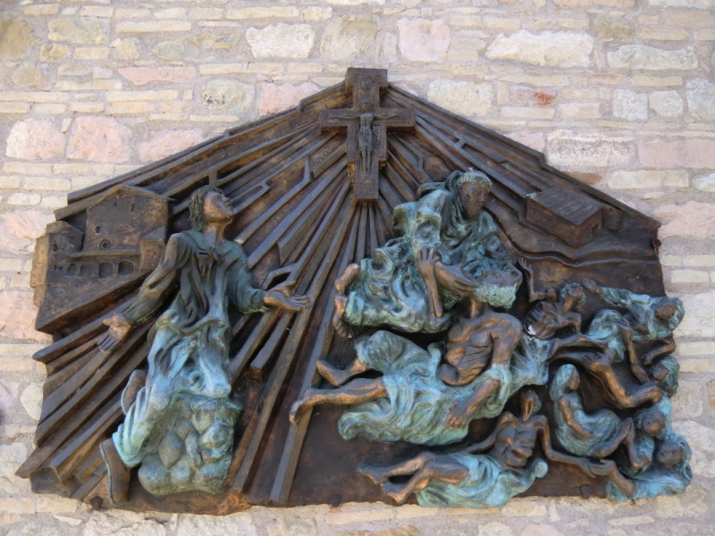 Silvio Amelio: „Bekehrung des Franziskus - Ja zu Christus und zu den Gefährten”; links im Hintergrund die Kirche des Klosters San Damiano, der Ort des Geschehens; Bronzerelief, 2006, am Santuario Santuario Rivotorto